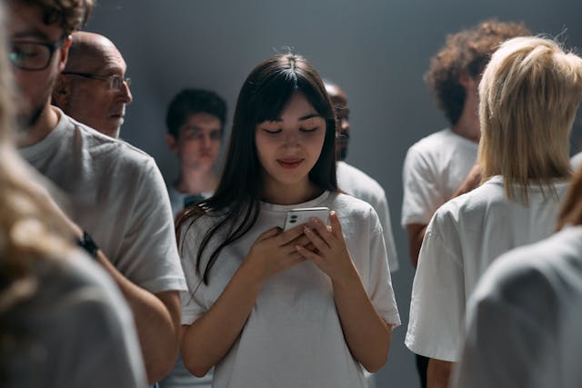 Young woman in a white t-shirt standing among a group of people, holding a smartphone and looking at the screen. The group is gathered in a softly lit indoor space, all wearing white t-shirts, creating a neutral and contemplative atmosphere.